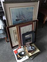 Eight framed art pieces and frames arranged leaning on furniture and on the floor, showing variety of frame sizes and art themes including ship, historical prints, and other illustrations.