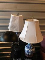 All three lamps together on black table, showing two Asian-style blue and white porcelain lamps with beige shades and one modern black metal lamp with a dome-shaped shade.