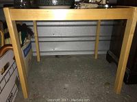 Light wood rectangular table with straight legs on cement floor, showing minor surface scratches