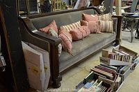 Front angle of vintage Chesterfield sofa with multiple decorative pillows on it, wooden arm with carved detailing visible, fabric faded with signs of wear and staining.