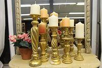 Full view of 9 gold decorative candle stands with various white and peach pillar candles on a yellow surface, next to an artificial plant with pink flowers in terracotta pot.