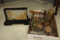 Lot of brass, metal and wooden decor items including a decorative Asian carved wooden frame on stand, brass figurines, horseshoe, stirrups, bangle, salt and pepper shakers, and a wooden hide hammer in a box.