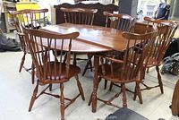 Full view of drop leaf dining table with 6 wooden chairs around it. The table is medium brown wood with two leaves and turned legs. Chairs are wooden spindled backs with turned legs.