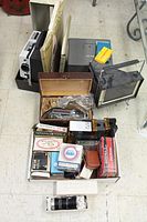 Overview photo of lot showing Brumberger metal reel case with reels, Bell and Howell video camera in case, various film accessories, film splicers, flash bulbs, and projection lens.