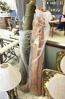 Four bolts of fabric shown upright. Largest is the green roll leaning on wall. Other smaller bolts include rusty orange color and a striped roll wrapped in plastic.