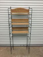 Full view of shelving unit standing open against a storage unit door, displaying three wicker shelves and steel frame with arched wicker top panel.