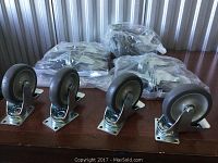 Four caster wheels on wooden surface in front of several bags containing additional wheels, showing metal mounting plates and swivel structure with brakes.