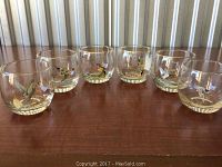 Set of six round glasses arranged in a row on wooden surface, each with hand-painted American duck motifs and a gold rim.