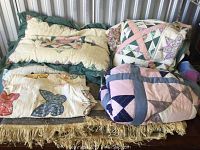 Lot containing three quilts stacked, two pillows resting on top, and a fringed woven rug underneath.