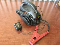 JNC300XL jump starter with attached jumper clamps and plug-in charger on a wooden surface.