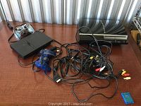 Photo showing PlayStation 3 console, PlayStation 2 console, two wired controllers and several cords on a wooden surface.