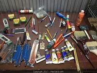 Full view of the art supplies spread on a table showing pencils, paint tubes, brushes, charcoal, erasers, paint scrapers, and rollers in various condition.