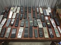 Wide view of multiple remote controls arranged on a table showing the variety in size, shape and button layout.