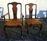 Two Queen Anne style oak dining chairs, one armchair and one side chair, shown outdoors on gravel with garage door background