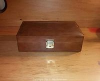 Front view of rectangular brown leather watch and jewelry case with silver-tone clasp lock on wooden surface.
