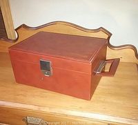 Side view of rectangular brown leather men's train case with metal lock on front and leather handle on top