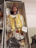 Porcelain doll laying in original box, dressed in Native American style clothing with a yellow fur vest and braided rope belt.