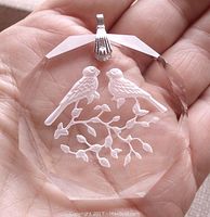 Close-up of clear crystal glass pendant held in hand showing carved love birds design and silver-tone bail.