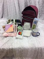 Full set of items including maroon backpack, plaid throw, candle, bento box, water bottle, note pads, pencil case, colored pencils, campmug, student cookbook and day planners arranged on white lace background.
