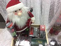 Photo showing a box containing a large plush Santa decoration, multiple boxes of Christmas balls in packaging, some labelled Krebs brand, two rolls of decorative ribbon, and the gold star tree topper visible.