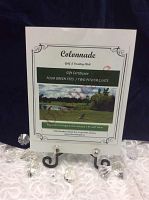 White paper gift certificate displayed on a black metal easel with lace tablecloth background. Certificate heading reads Colonnade Golf & Country Club with 'FOUR GREEN FEES / TWO POWER CARTS' and course image.