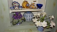 Full photo of shelf including vintage Asian peanut bowls, tea set with yellow camel teapot, lidded blue and white urn, cobalt blue glass bowl, and two flower arrangements.