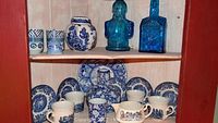 Photo showing various blue and white plates, mugs, pitchers, and two blue decorative glass bottles on wooden shelving.
