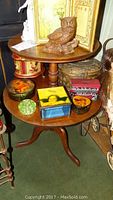 Photo of two-tier wood table with various decorative items including carved wooden owl figurine, framed artwork, metal tins with rooster and cherub designs, green glass flower frog, small blue glass box with cherub image, woven wicker basket, and a red metal tin with afternoon tea design on lower tier.