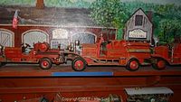 Three red reproduction antique toy fire trucks arranged side by side on a shelf against a painted backdrop.