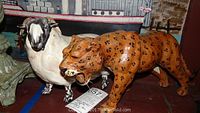 Photo of leather leopard figurine with black spots standing next to white and black porcelain ram figurine on table