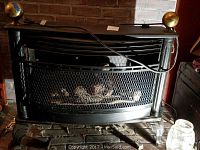 Front view of black metal propane heater with brass top finials and mesh safety screen showing faux logs inside, with electric cord on top.