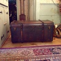 Front and side view of the antique wooden shipping trunk on carpet, showing metal bands and latches, and distressed paint finish.