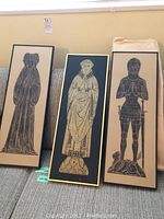 Three framed brass rubbings side-by-side showing full figures in medieval style on beige backgrounds.