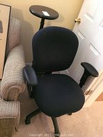Black adjustable office chair with armrests and a caster wheel base shown next to a couch and door, seat and backrest padded and covered in fabric.