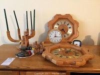 Two octagonal oak wall clocks, one standing up and one lying flat, positioned behind a group of decorative wooden items including a wooden candle holder and bird figurines on a wooden surface.