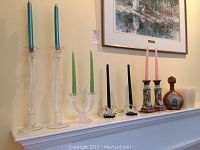 Wide shot of all candle holders arranged on mantel including 2 tall glass candle sticks with blue candles, double glass green candle holder, black candles in small holders, 2 ceramic floral holders with pink candles, and round leather covered decanter.