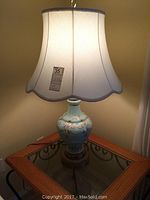Light blue glass base table lamp with cream-colored scalloped fabric lampshade placed on glass and wooden table, illuminated to show lampshade shape and size.