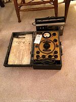 Overview of tube tester open showing dial, sockets, cord, and instruction sheet inside case, with Sony radio behind.