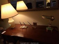 Desk with a glass-bodied table lamp, brass bankers lamp, Seiko clock, and four pen holders arranged on top.