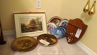 Photo showing eight decorative plates arranged on carpet against wall, including some blue and white plates, two brown-toned plates with scenes, a framed picture, wooden spoon rack on the right, and place mats.
