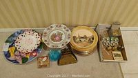 Assorted decorative plates including blue glass dish, floral ceramic plate, gold charger plates with village scenes, and boxes containing napkin rings and knife rests.