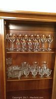 Shelved display of nine crystal wine glasses with gold rims on top shelf, eight sherbet glasses below, and a crystal bowl and vase on lower shelf.