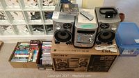 Sony compact stereo system with two speakers, remote control on top, and box of CDs and DVDs beside it, showing full lot contents.