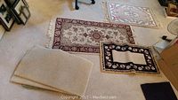 Overview of multiple rugs and mats laid on carpeted floor, showing different sizes and types including folded beige mat and black floral rug
