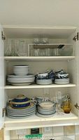 Inside kitchen cabinet showing stacked plates, cups, saucers, bowls and some glassware including clear drinking glasses arranged on shelves.