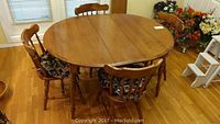 Photo showing oval wood dining table with two leaves and four cushioned wooden chairs around table on hardwood floor.