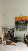 Framed artwork, woven wall hanging with house and trees, dark carved stone seal sculpture, cat chime and wooden chime parts on carpeted floor