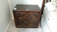 Left side photo of one wood night stand with two drawers and intricate carved fronts in dark wood finish near bed.