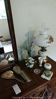 Full view of dressing table set including wooden mirror, hairbrush, comb, artificial flower vase, lidded bowl, ring holder, floral cup, floral cloth, and the electric lamp on a wooden surface