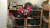 Photo showing wooden shelves holding various Christmas decorations including a plush reindeer head wall hanging with red polka dot antlers, an artificial wreath with red bow, and stacked holiday boxes